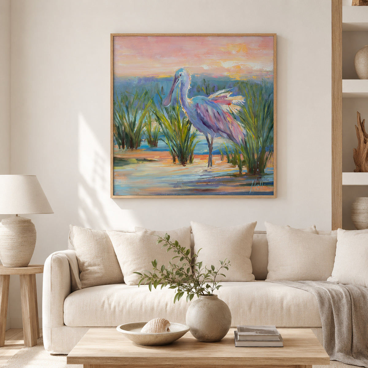 A framed painting of a pink spoonbill bird standing in reeds under a pink and orange sky, displayed on a light beige wall above a cream sofa in a modern living room setting.