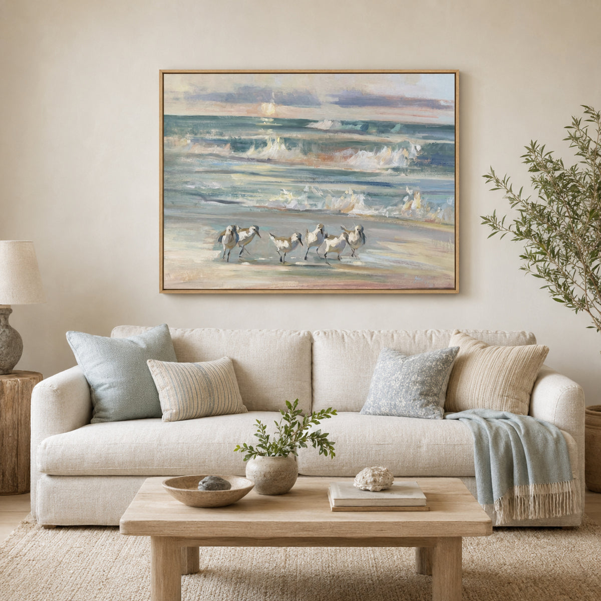 A framed beach painting hanging on a beige wall above a light-colored sofa in a styled living room. The artwork depicts small birds walking on a sandy shore with ocean waves behind them in soft blue, beige, and gray tones.