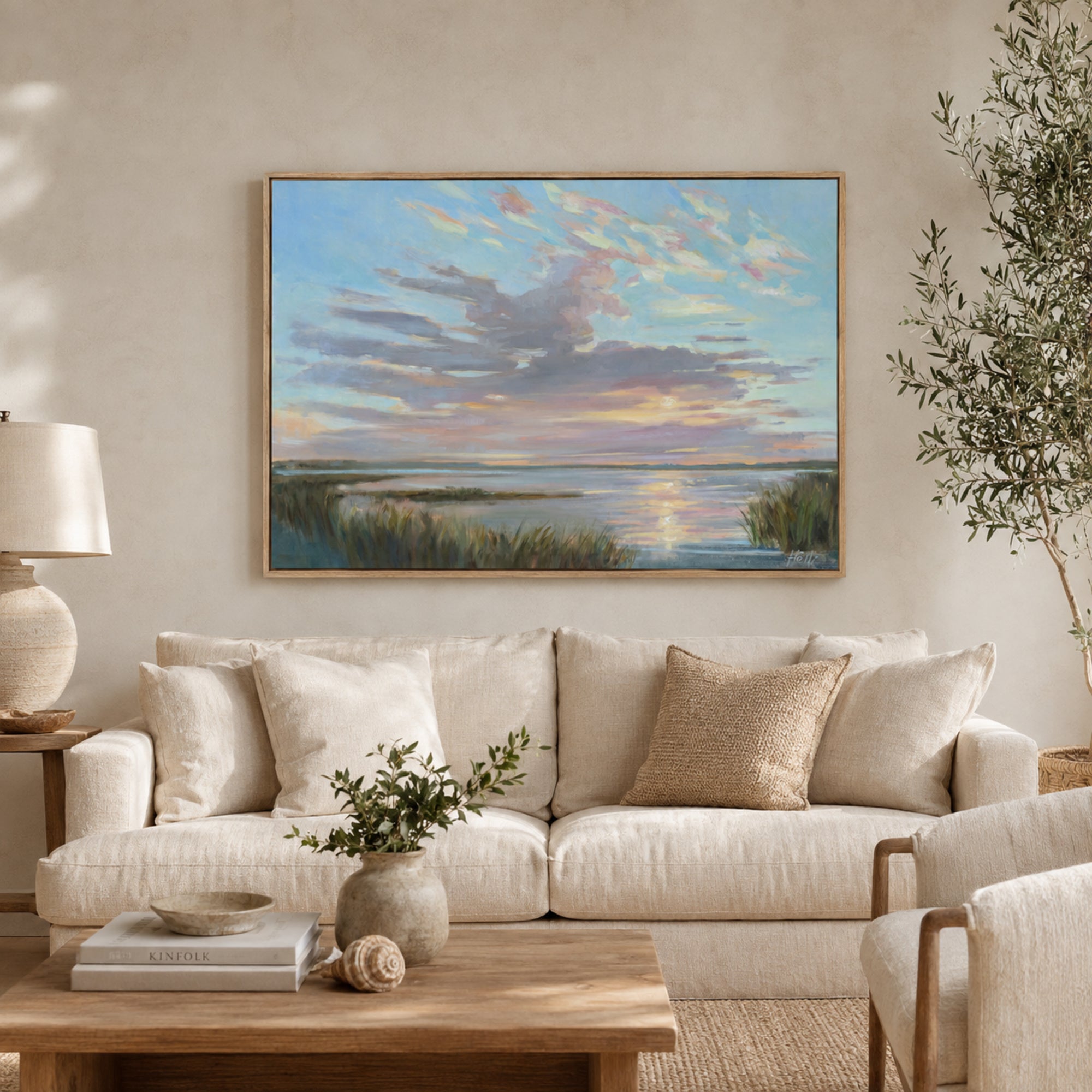 A framed landscape painting of a sunset over water with dramatic clouds hangs on a beige wall above a cream-colored sofa in a styled living room.