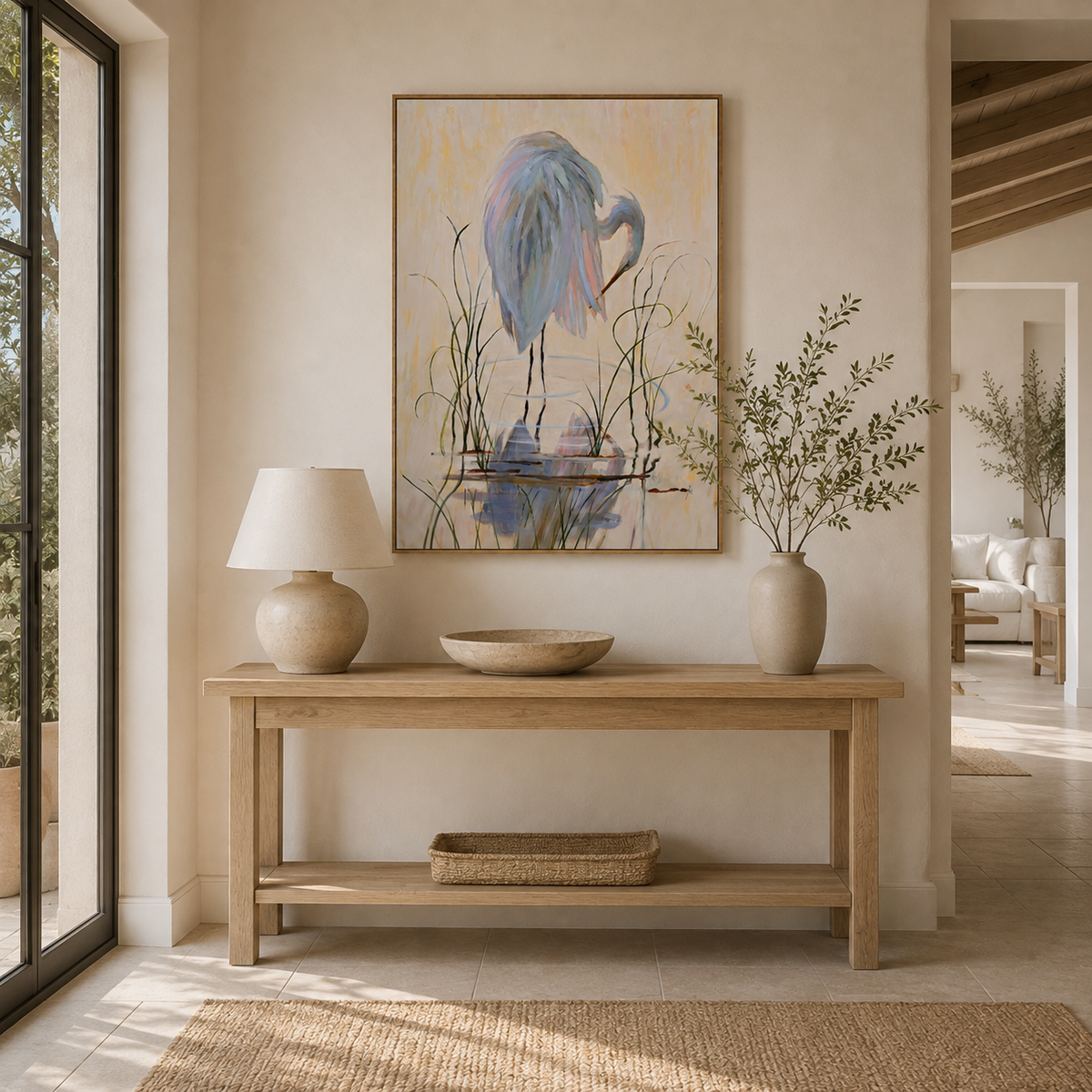 A framed vertical painting of a blue heron standing in reeds hangs on a beige wall above a light wood console table in a bright, modern interior.