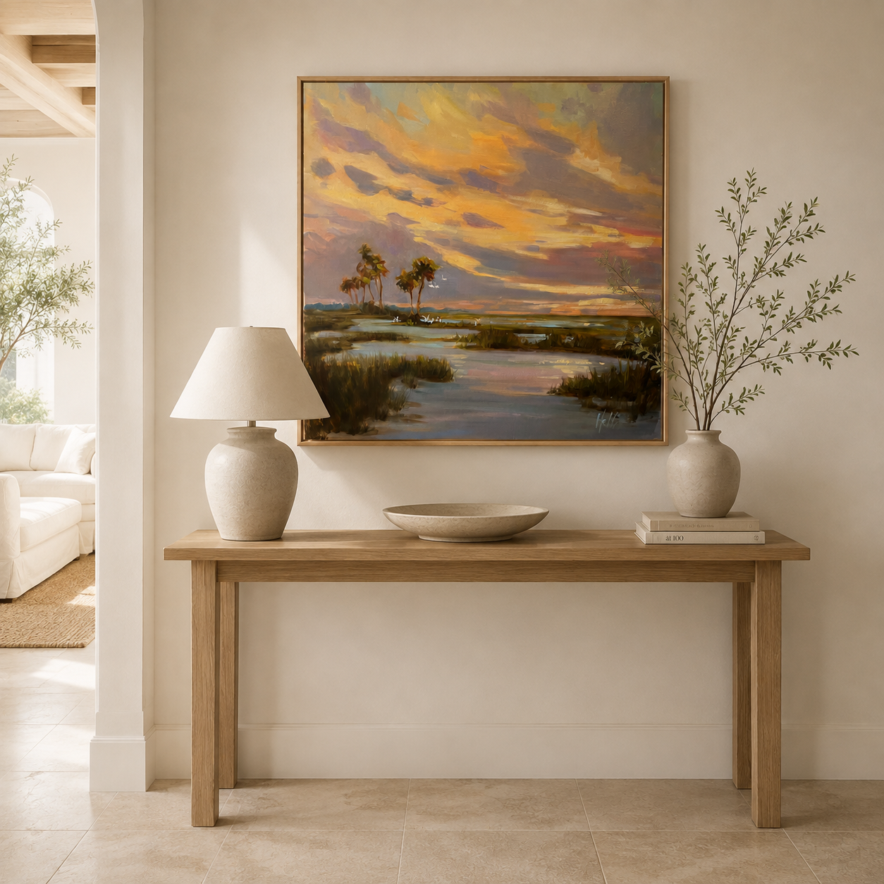 A framed landscape painting of a sunset over marshlands with palm trees hangs on a beige wall above a light wood console table. The scene includes a table lamp on the left, a shallow bowl in the center, and a vase with branches on the right.