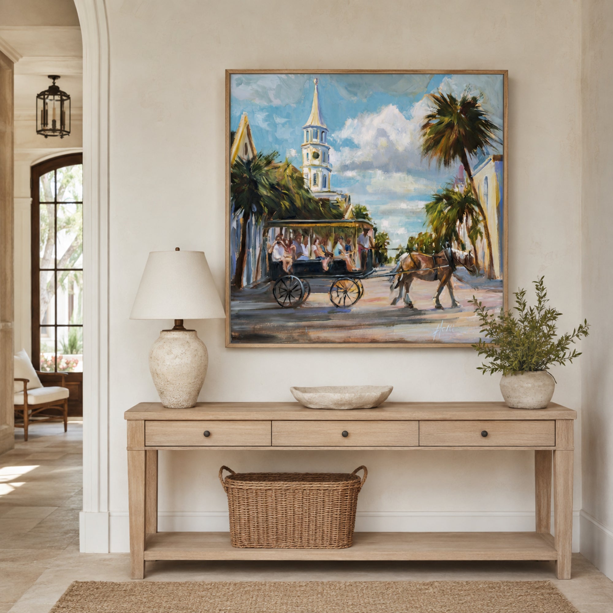 A framed painting of a horse-drawn carriage scene with palm trees and a church steeple, displayed on a beige wall above a light wood console table in a styled interior room.