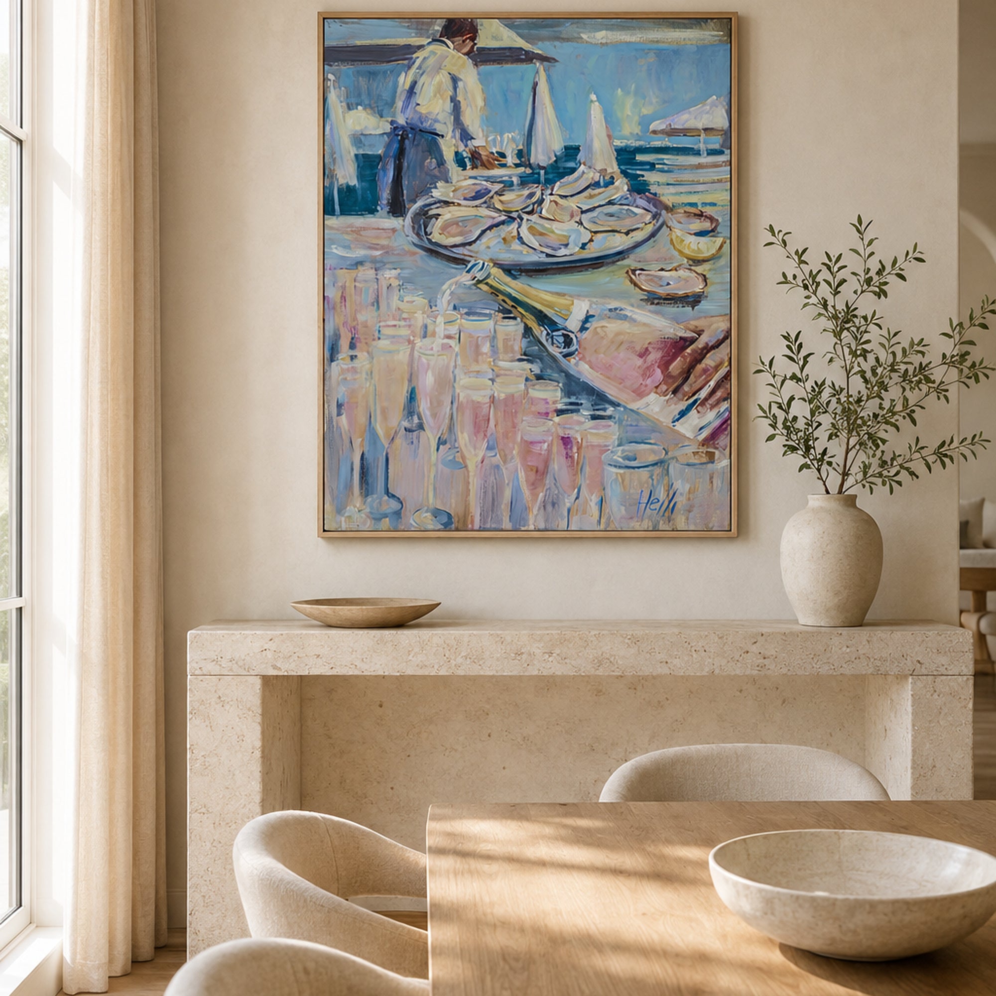A framed painting of a coastal dining scene hangs on a beige wall above a stone console table in a bright, modern dining room. The artwork features blue, pink, and white tones with figures, plates, and reflections, and is styled in a warm, minimalist interior with a vase of greenery and wooden dining chairs.