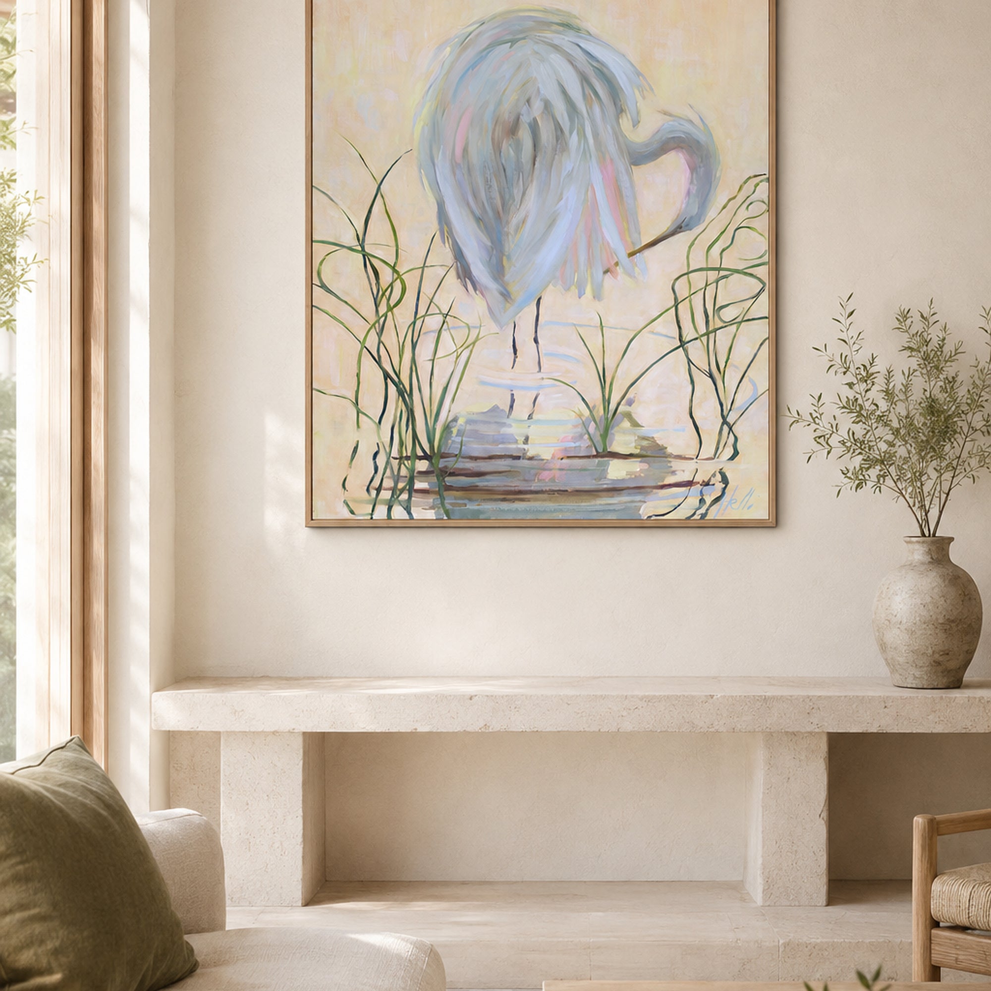 A framed abstract painting of a heron and reeds hangs on a beige wall above a stone bench in a softly lit room. The artwork features soft blue, gray, green, and cream tones, and the scene includes a vase with dried stems and part of a sofa with a green cushion.