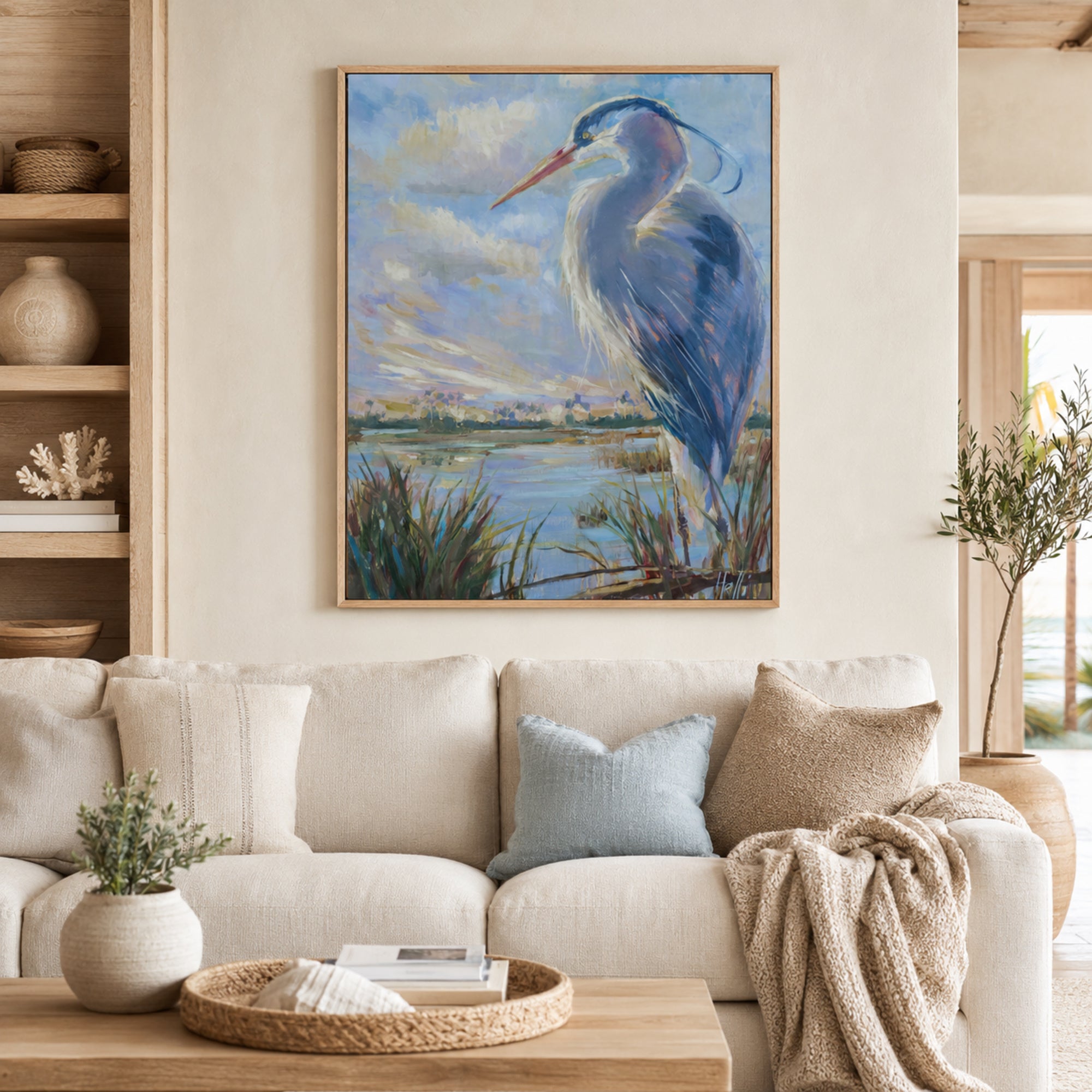 A framed vertical canvas print of a blue heron standing by a lake, displayed on a beige wall above a cream-colored sofa in a styled living room setting.