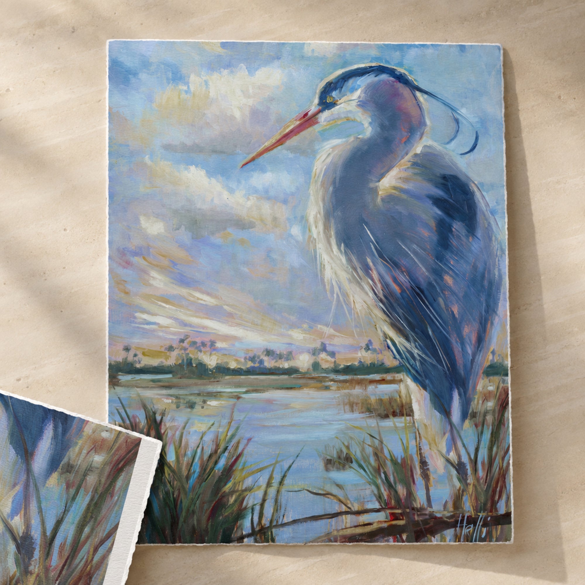A square canvas painting of a blue heron standing in a marshy landscape under a cloudy sky, displayed on a light wood surface with another cropped view of the artwork partially visible in the lower left corner.