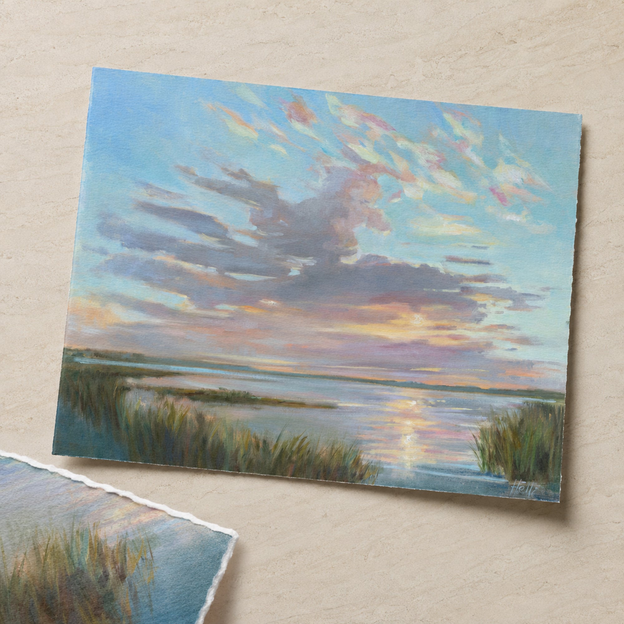 A square art print or painting depicting a calm lake or marsh scene at sunset, with reeds in the foreground and a blue sky filled with soft pink, purple, and yellow clouds. The print is photographed on a light beige textured surface, with part of another torn paper corner visible in the lower left.