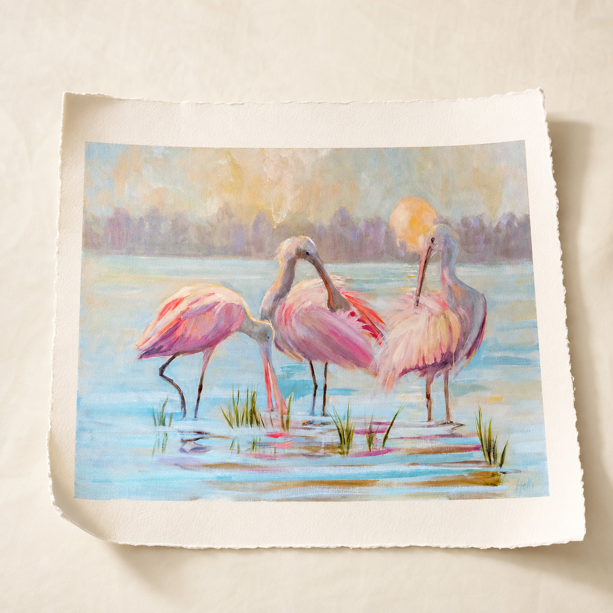 A square art print featuring a pastel watercolor painting of three pink spoonbills standing in shallow blue water, with a soft yellow sun and blurred trees in the background, displayed on a light beige textured surface.
