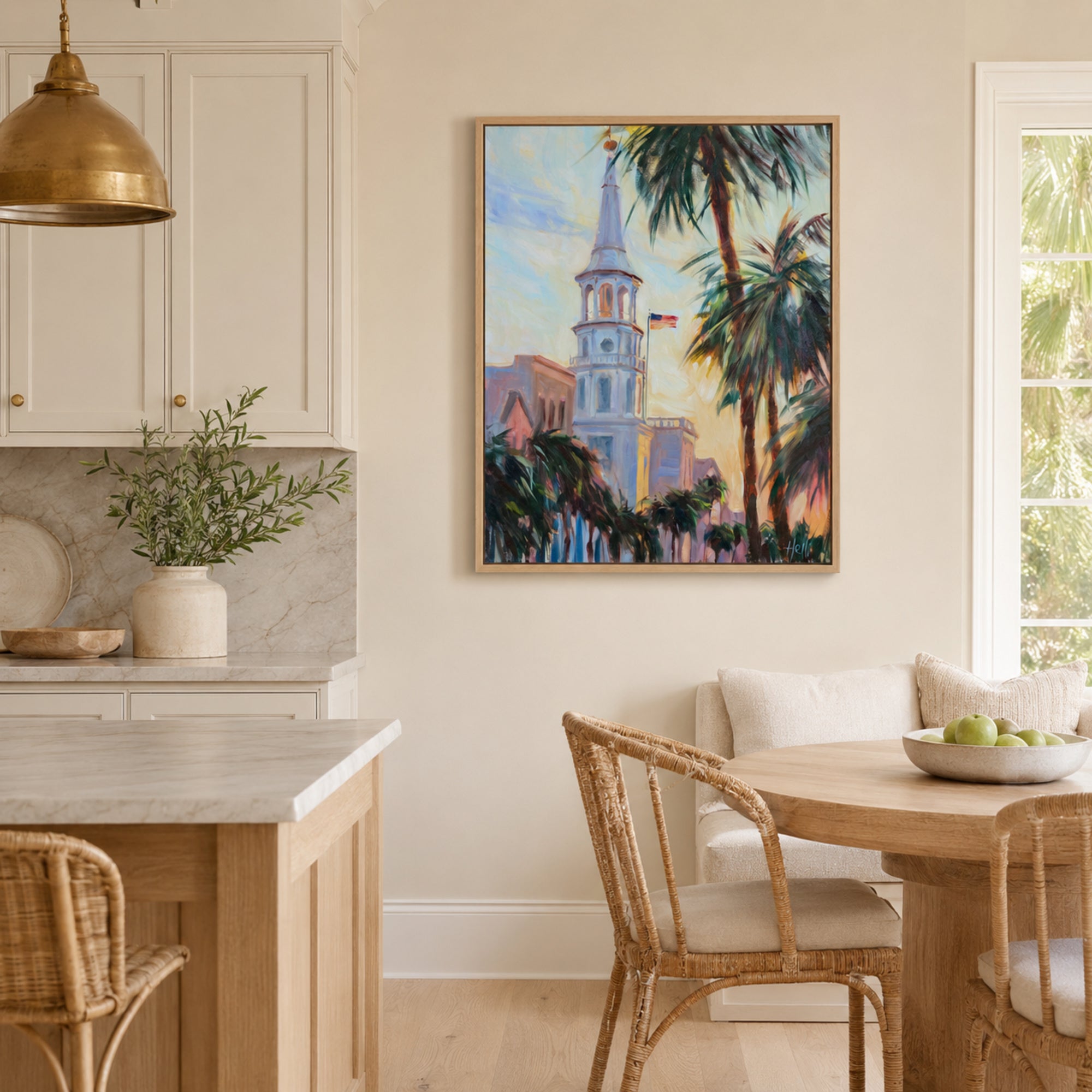 A framed colorful painting of a church tower and palm trees hangs on a beige wall in a bright kitchen and dining room setting.
