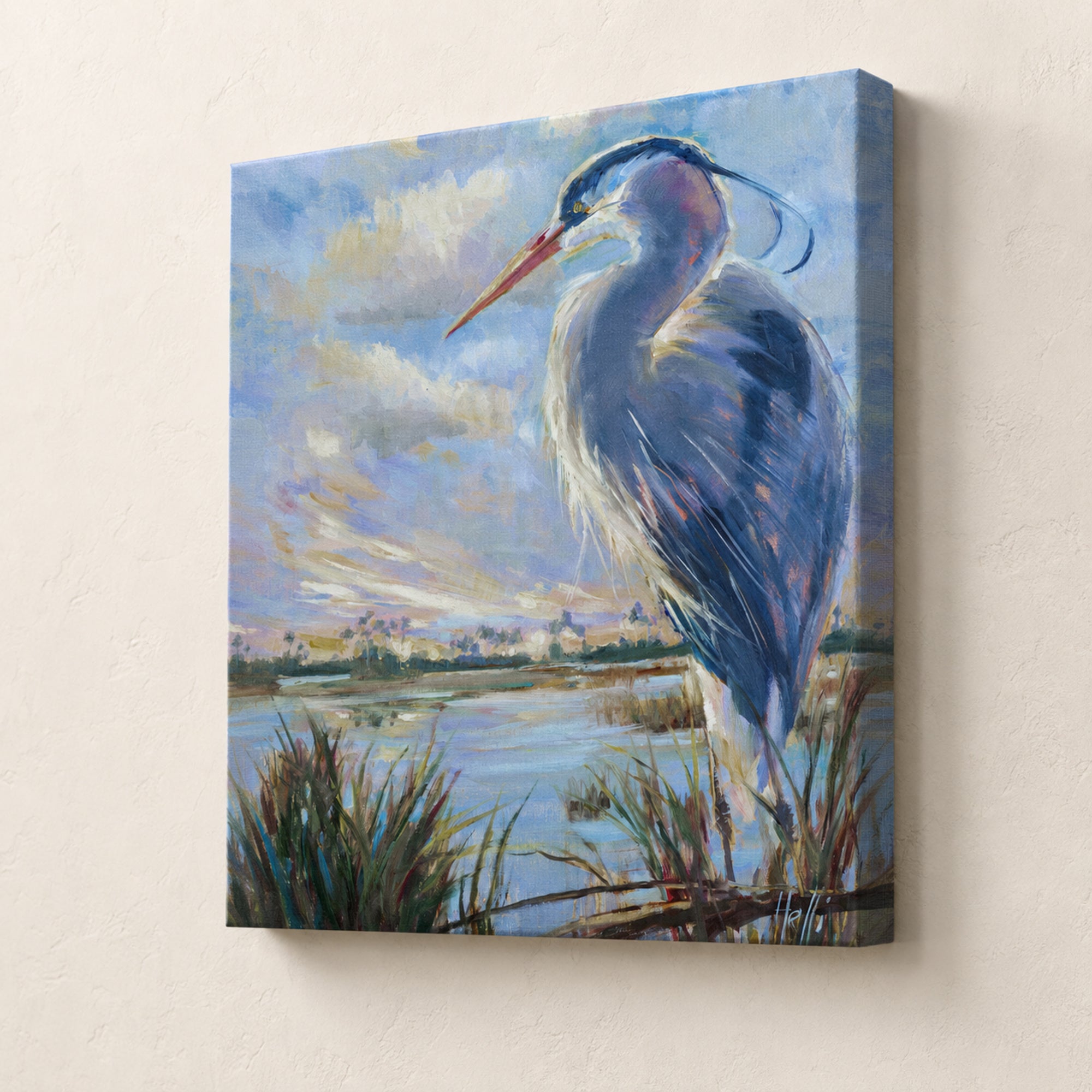 A square canvas wall art print depicting a blue heron standing in shallow water under a cloudy sky, shown hanging on a light beige textured wall.