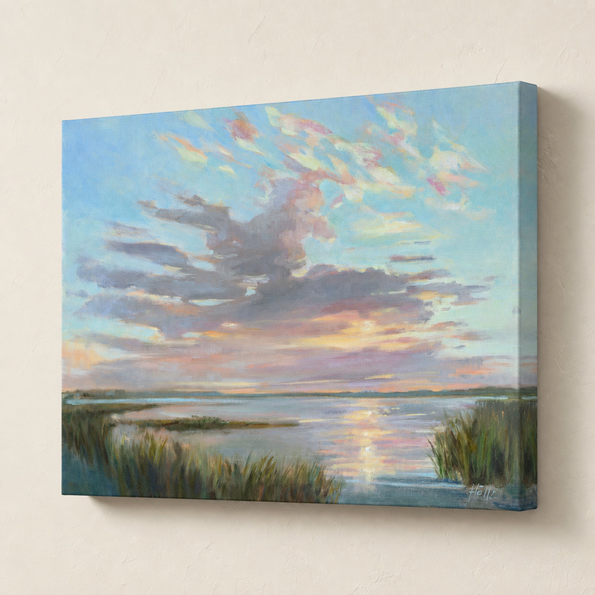 A square canvas wall art print depicting a calm lake or marsh scene at sunset, with blue sky, soft pink and peach clouds, reflective water, and dark reeds in the foreground. The canvas is shown hanging against a light beige wall.