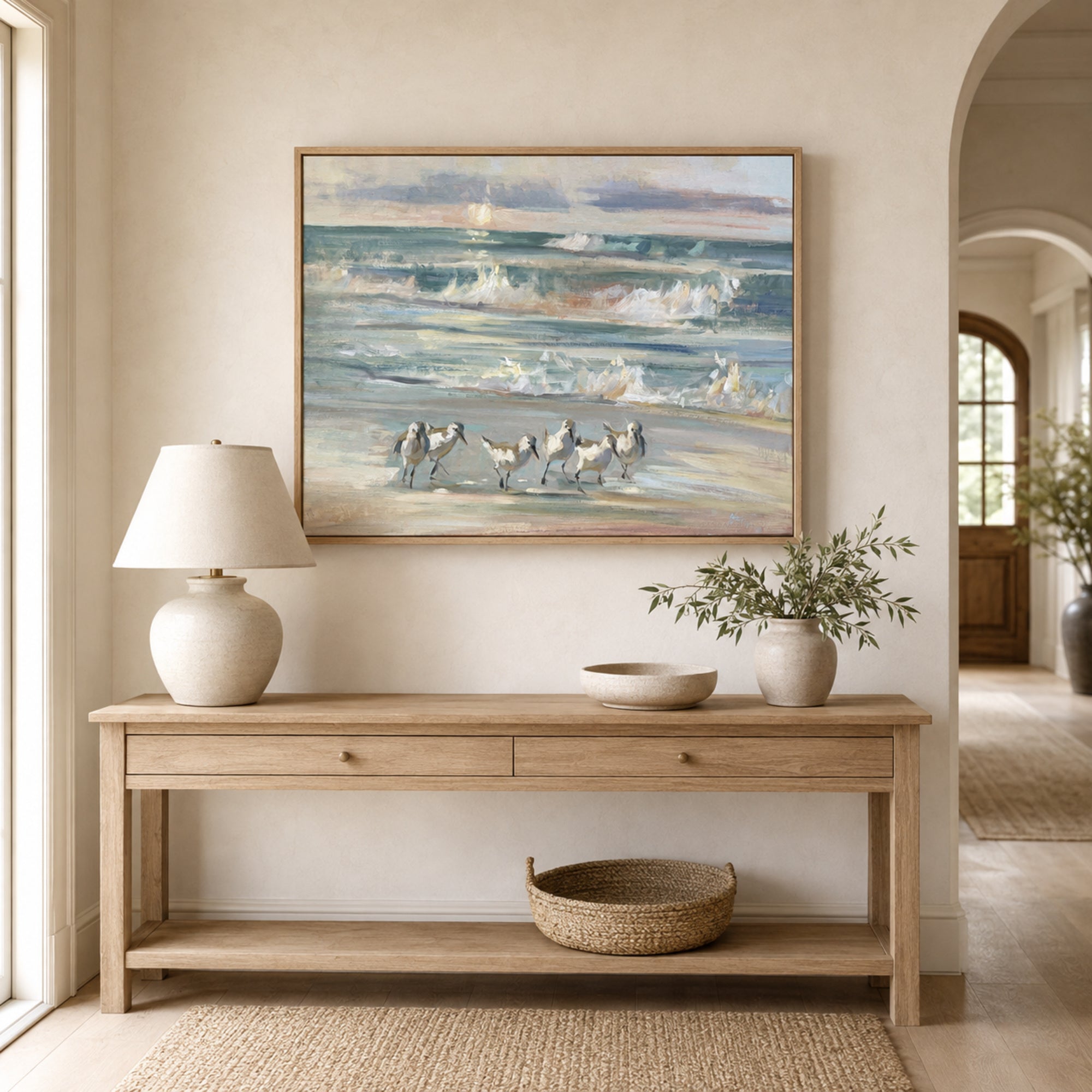 A framed beach painting hangs on a beige wall above a light wood console table in a styled interior. The artwork depicts waves, a sailboat, and small figures on a sandy shore.