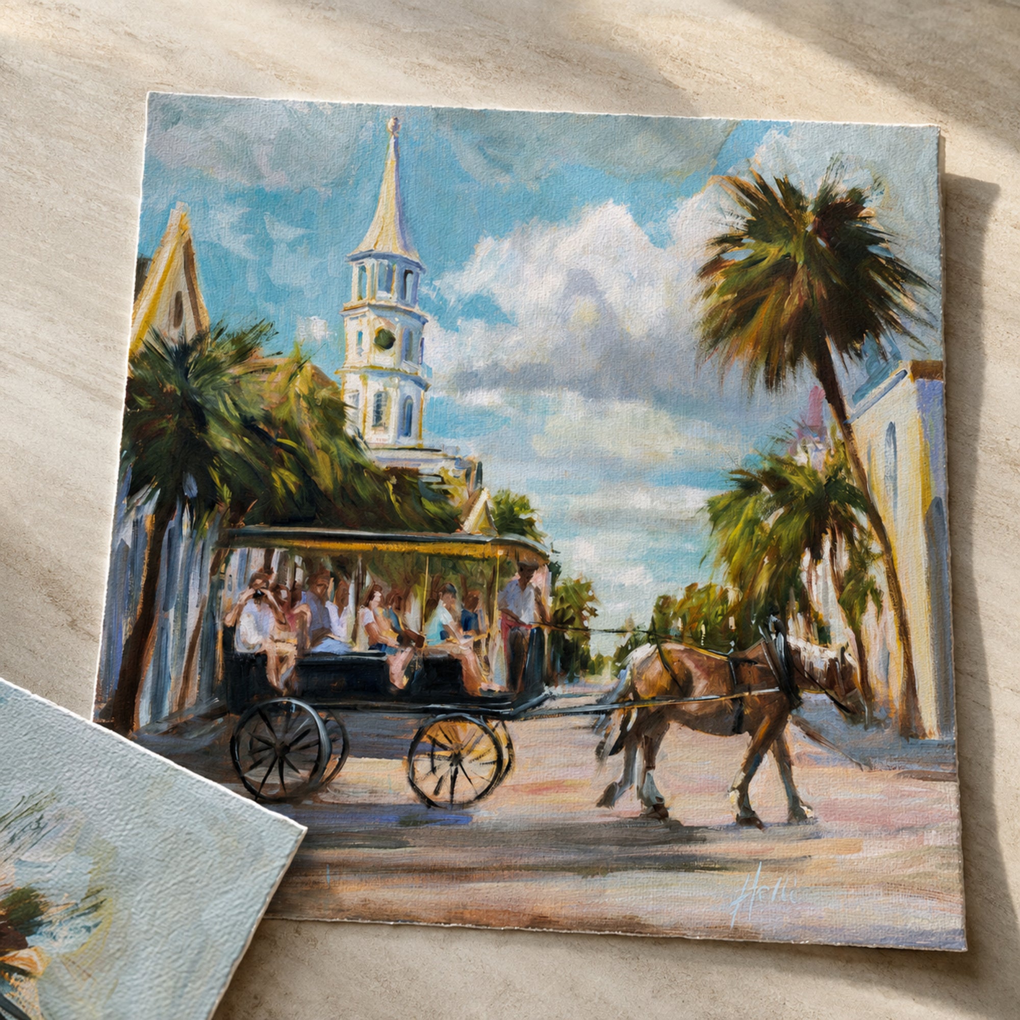 A square canvas painting of a coastal street scene with a horse-drawn carriage, palm trees, a church steeple, and a blue sky, displayed on a light wood surface.