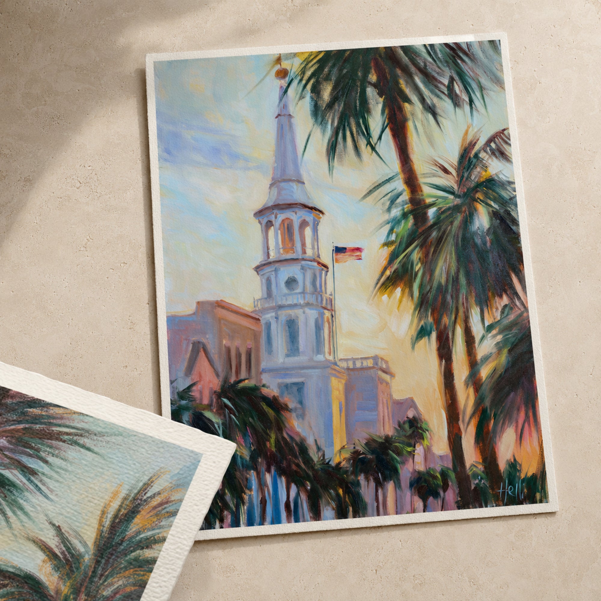 A square art print featuring an impressionistic painting of a white church steeple with palm trees and a warm sunset sky, displayed on a textured beige surface with another cropped print partially visible in the lower left.