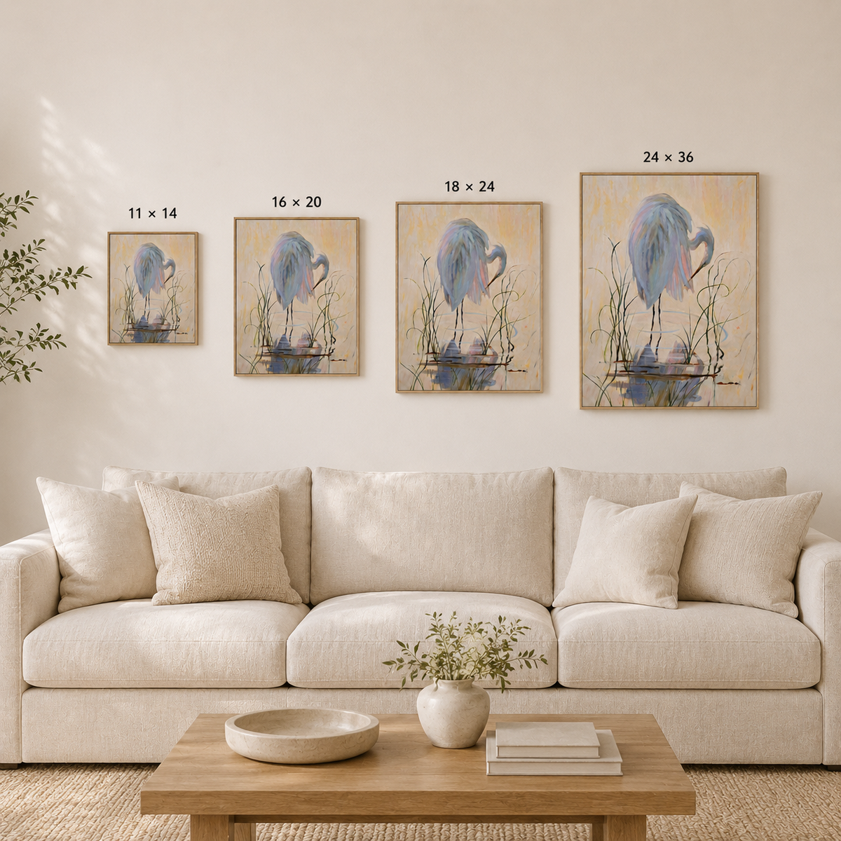 A staged living room showing four framed bird wall art prints in different sizes mounted above a cream sofa. The prints depict a blue heron standing in reeds on a warm beige background, with size labels above each frame.